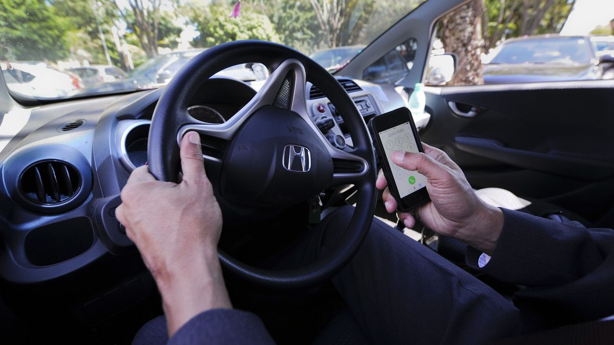 Motorista segura o celular com uma das mãos e o volante com a outra, dentro de um carro