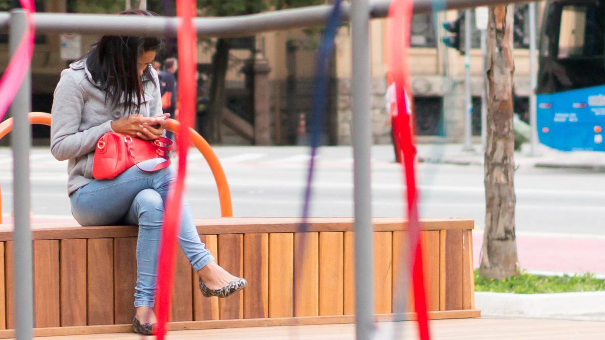 Mulher usa o celular sentada num banco no centro de São Paulo, com ônibus ao fundo