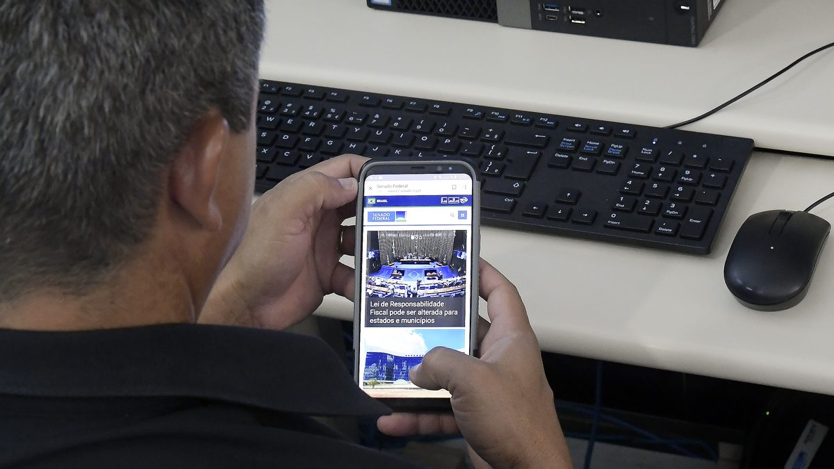 Homem consulta um site no celular sentado diante do computador, no trabalho